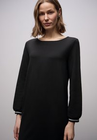 Czarna sukienka z długimi rękawami, okrągłym dekoltem i dopasowanym stanem. Z białymi paskami ozdobnymi na mankietach rękawów. Gładka faktura tkaniny.