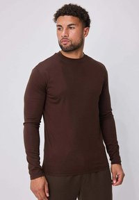 T-shirt marron à manches longues avec col rond, fabriqué en tissu lisse, coupe ajustée et conception simple. Aucun motif ou accent visible.