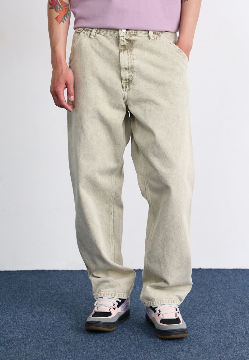 Hellbeige, lockere Jeans mit leicht strukturiertem Finish, ausgestattet mit Fronttaschen und einem Knopfverschluss, kombiniert mit mehrfarbigen Turnschuhen.