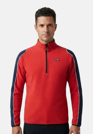Uomo che indossa un pile rosso con zip a quarti, strisce blu navy e piping bianco lungo le maniche, in piedi davanti a uno sfondo semplice.