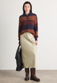 Pull en tricot rayé rouille, marine et marron, avec un col et trois boutons. Jupe midi en satin olive, associée à des bottes marron.