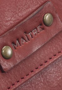 Roter Lederanhänger mit eingeprägtem Text "MAÎTRE", ausgestattet mit zwei messingfarbenen Nieten und rosa Ziernähten entlang der Kanten. Sichtbare strukturierte Oberflächendetails.