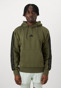 Sweatshirt verde oliva com um bolso frontal, design em preto com três riscas nas mangas e um pequeno logótipo preto no peito. Textura de tecido suave.
