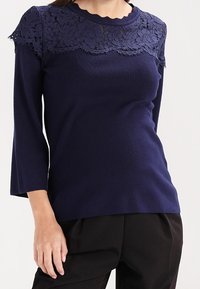 Top de manga larga azul marino con detalles de encaje en la parte superior del pecho y los hombros, combinado con pantalones negros.