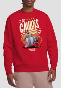 Red sweatshirt featuring a cartoon graphic with two characters, a dog, and text reading "Les Gaulois." Ribbed cuffs and hem. Soft fabric texture.