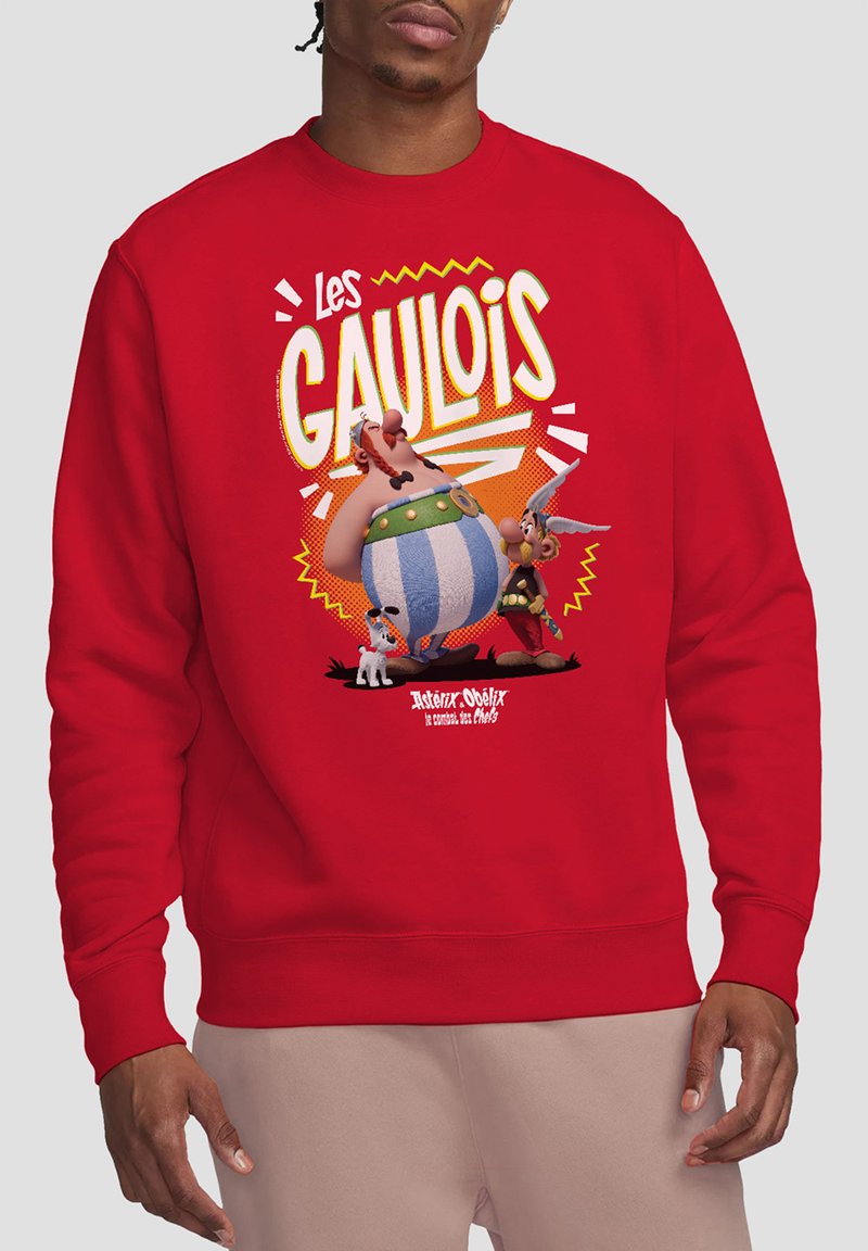 Red sweatshirt featuring a cartoon graphic with two characters, a dog, and text reading "Les Gaulois." Ribbed cuffs and hem. Soft fabric texture.