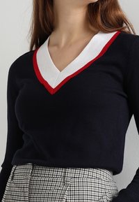 Jersey azul marino con un cuello en V blanco y rojo, con un diseño ajustado. Combinado con una falda de cintura alta de patrón a cuadros.