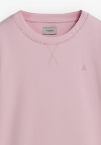 Suéter rosa claro con un cuello redondo acanalado, que presenta un pequeño logo bordado en el lado izquierdo del pecho y detalles de punto de cruz.