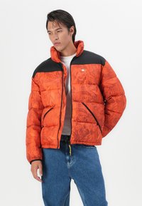 Doudoune de couleur orange vif avec un motif floral, des accents noirs sur les épaules, fermeture éclair et poches latérales. Portée par-dessus une chemise grise.