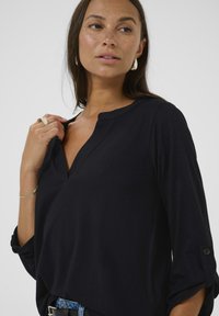 Chemise noire à manches longues avec un décolleté en V, fabriquée en tissu doux. Présente des manches roulées avec des accents de boutons et une coupe décontractée.