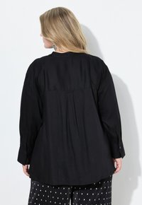 Camicia nera a maniche lunghe con piega sul retro, dotata di polsini con bottoni. Il tessuto sembra liscio con una vestibilità comoda, abbinata a pantaloni a pois.