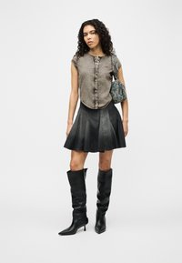 Γυναίκα με μπουκλωτά μαλλιά που φοράει γκρι αμάνικο τοπ με κουμπιά, μαύρη φούστα με καμπάνα, μαύρες μπότες μέχρι το γόνατο και κρατάει μια μπλε καπιτονέ τσάντα ώμου.