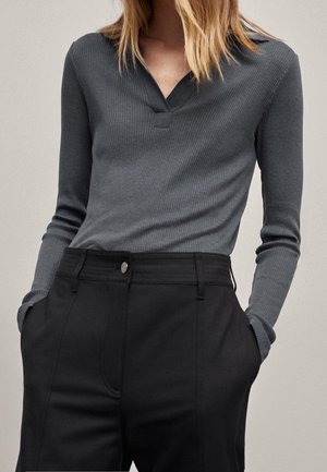 Femme portant un haut à col en V gris anthracite à manches longues côtelé, rentré dans un pantalon noir taille haute, les mains dans les poches.