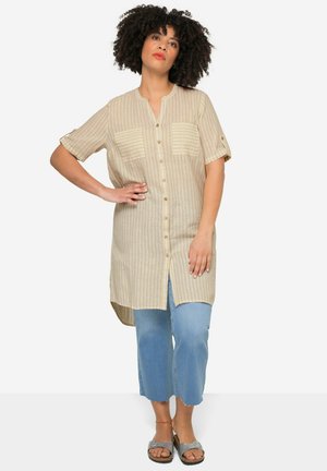 Beige gestreepte shirtjurk met korte mouwen, knoopsluiting aan de voorkant, twee zakken op de borst, gebogen zoom, gedragen over lichtblauwe cropped jeans en sandalen.