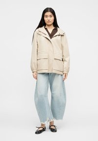 Kobieta w beżowej kurtce z kapturem, ciemnobrązowej koszuli, jasnoniebieskich szerokich dżinsach z postrzępionymi krawędziami i czarnych balerinach, stojąca na białym tle.