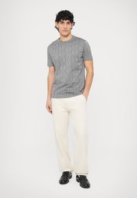 Szary t-shirt z krótkim rękawem o fakturowanym wzorze, zestawiony z kremowymi spodniami o prostych nogawkach i czarnymi butami. Jednolite tło.