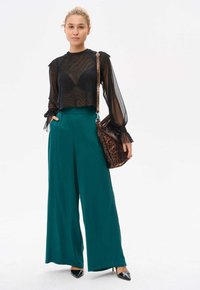 Czarna, przezroczysta bluzka z długim rękawem i falbaniastymi mankietami, zestawiona z turkusowymi spodniami o szerokich nogawkach. Stylizację dopełniają torebka w panterkę i czarne szpilki.
