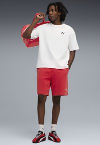Hvid kortærmet skjorte med sort logo, røde shorts, røde og sorte sneakers, holder en blank rød taske. Simpelt design, sporty look.