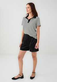 Polo a righe bianco e nero con colletto nero, abbinato a pantaloni corti in satin neri e ballerine nere. Texture liscia in tutto il capo.