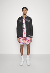 Chaqueta negra de botones con "NEW YORK" impreso en el pecho, combinada con una camisa de patrón de manchas coloridas y pantalones cortos a juego. Zapatillas blancas.