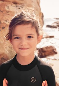 Black and orange wetsuit featuring a circular logo. The child has medium brown hair and stands in front of rocky beach scenery.