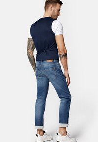 Granatowa kamizelka z tkaniny nałożona na białą koszulkę, zestawiona z wyblakłymi niebieskimi dżinsami i białymi sneakersami. Posiada podwinięte rękawy i subtelny detal w postaci wstążki z tyłu.