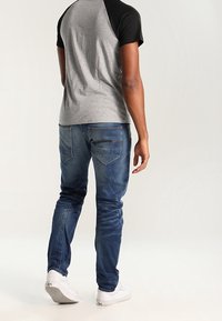 Czarna i szara koszulka raglanowa w połączeniu z niebieskimi dżinsami. Białe trampki. Widok z tyłu, ukazujący dopasowanie i detale projektu, takie jak umiejscowienie kieszeni.