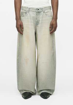 Personne portant un jean gris délavé à jambes larges avec coutures visibles, poches avant, fermeture par boutons et chaussures noires à bouts pointus sur fond blanc.