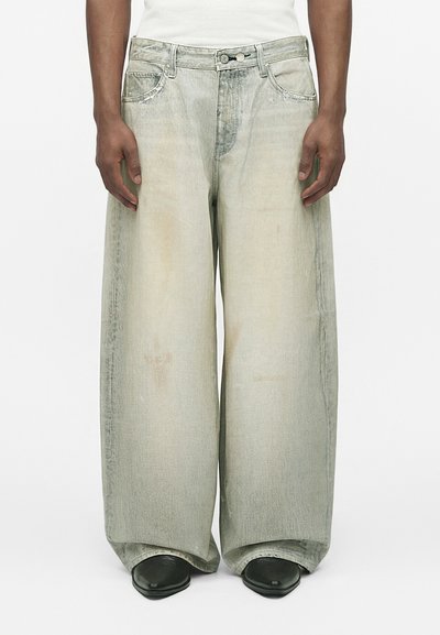 Person wearing wide-leg, faded gray jeans with visible seams, front pockets, button closure, and black pointed-toe shoes on a white background.