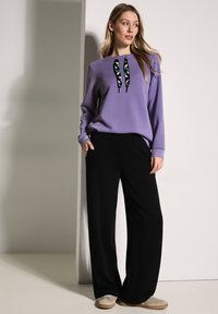 Felpa viola con lacci decorati, pantaloni neri a gamba larga e sneaker beige. Tessuto morbido e vestibilità rilassata. Posizione in piedi.