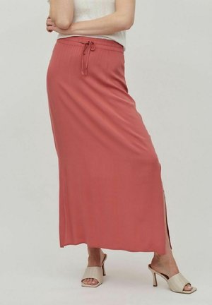 Kvinde iført et langt, støvet rosa skørt med snøre i taljen og beige åbne hæle, stående med armene krydsede.