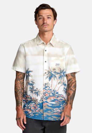 Uomo con tatuaggi che indossa una camicia bianca a maniche corte con motivo di palme e fiori blu e rosa, in piedi davanti a uno sfondo uniforme.