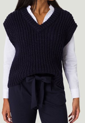 Vrouw die een wit shirt met lange mouwen draagt onder een navyblauwe grofgebreide mouwloze V-hals trui met bijpassende navyblauwe broek die is vastgebonden met een stoffen riem.