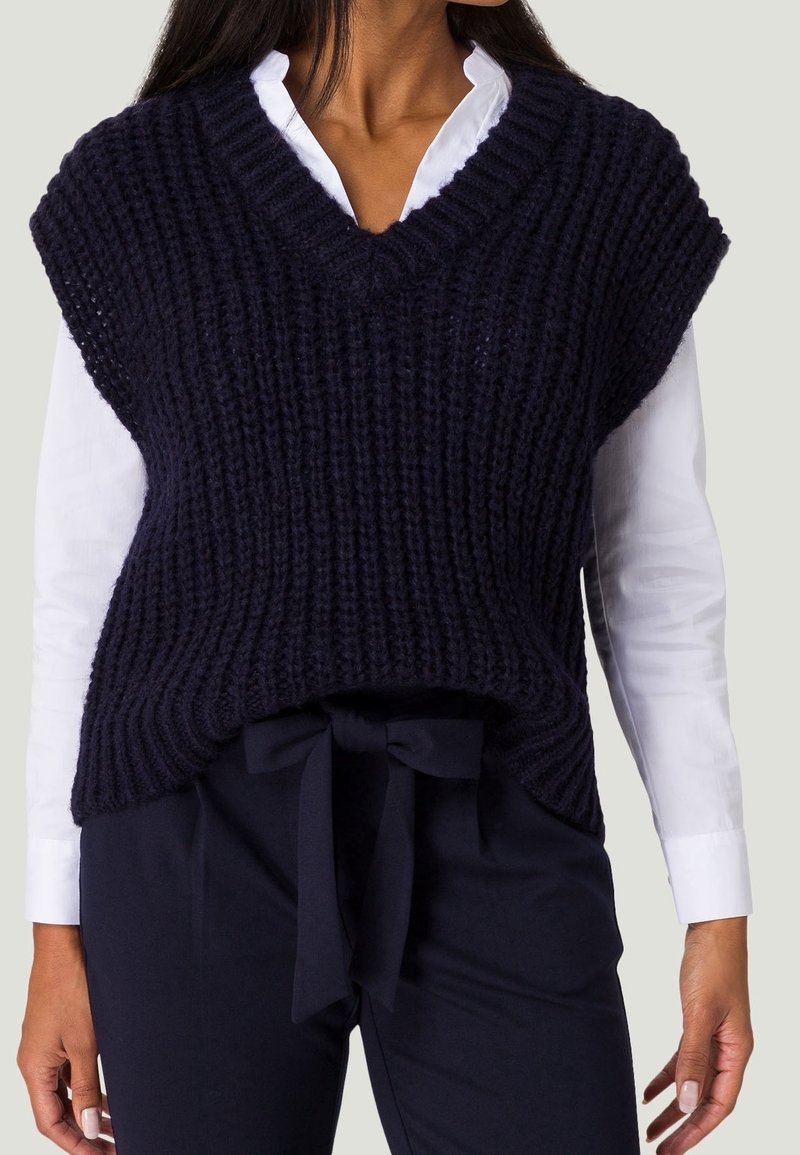 Femme portant une chemise blanche à manches longues sous un pull sans manches à col en V en grosse maille bleu marine, associé à un pantalon bleu marine noué avec une ceinture en tissu.