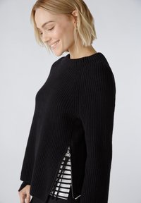 Schwarzer Strickpulli mit geribbelter Textur, Rundhalsausschnitt, seitlichen Reißverschlüssen und einem gestreiften Hemd, das am Saum hervorschaut. Lange Ärmel.