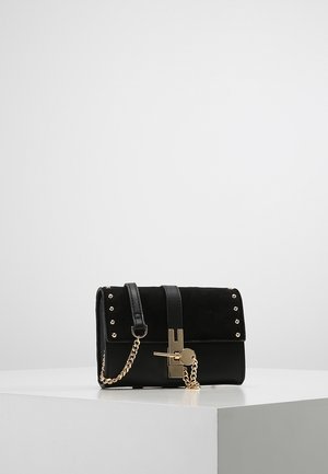 Pochette en cuir et daim noir avec clous dorés, bandoulière chaîne et détail de fermoir doré reposant sur un piédestal géométrique blanc.