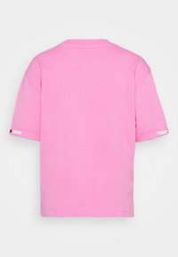 Camiseta corta de manga rosa sólido con el texto y el logotipo de "TOMMY JEANS" en los dobladillos de las mangas, mostrada desde la parte de atrás.