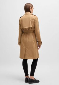 Langer beiger Trenchcoat mit Schulterklappen, tailliertem Gürtel und ventiliertem Rücken, kombiniert mit schwarzen Leggings und Schuhen.