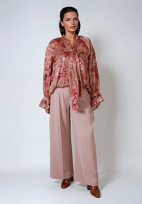 Luftige, durchsichtige Bluse in Rosa mit Blumenmuster und Bindedetail, kombiniert mit weit geschnittenen, hellrosa Hosen und braunen Stiefeletten mit Absatz.