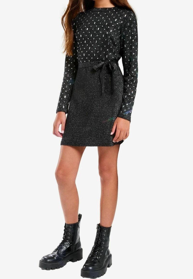 Jeune femme portant une robe noire à manches longues avec des pois scintillants et une ceinture à la taille, associée à des bottes de combat noires à lacets.