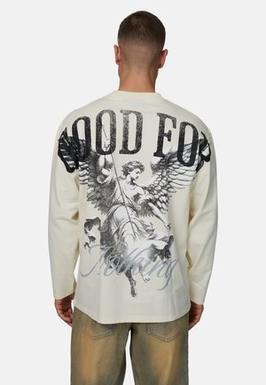 Kremowa koszula z długim rękawem z dużym czarnym nadrukiem anioła z skrzydłami, z napisem "GOOD FOR NOTHING" umieszczonym nad i pod grafiką.