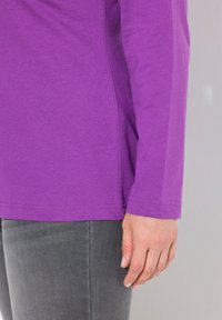 Lila Langarmshirt aus Baumwolle, mit rundem Halsausschnitt und dezenten Steppdetails, kombiniert mit grauen, taillierten Hosen.