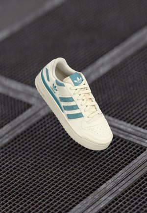 Valkoinen ja turkoosi Adidas-tennari kelluu mustan metalliverkkopinnan yläpuolella, näyttää sivu- ja yläkuvat kengännauhat sidottuina.