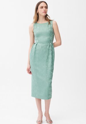 Kobieta ubrana w sukienkę midi z jacquardu w jasnym zielonym kolorze w kwiaty, bez rękawów, z dopasowaną talią, oraz beżowe buty na obcasie z odkrytymi palcami, stojąca na białym tle.
