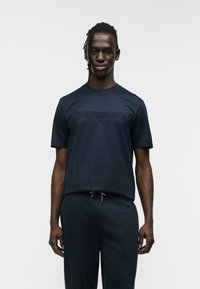 Μπλούζα από βαμβάκι σε ναυτικό μπλε χρώμα με κοντά μανίκια, που διαθέτει ανάγλυφο λογότυπο φτερού Aston Martin στο μπροστινό μέρος. Συνδυάζεται με παντελόνια σε σκούρο χρώμα που ταιριάζουν.