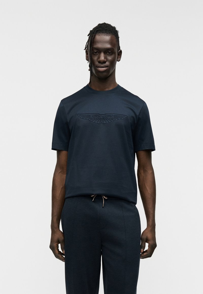 Μπλούζα από βαμβάκι σε ναυτικό μπλε χρώμα με κοντά μανίκια, που διαθέτει ανάγλυφο λογότυπο φτερού Aston Martin στο μπροστινό μέρος. Συνδυάζεται με παντελόνια σε σκούρο χρώμα που ταιριάζουν.