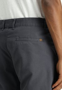 Donkergrijze stoffen broek met een gladde textuur, voorzien van een achterzak en een klein bruin logoaccent nabij de zak.