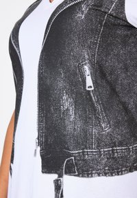 Camiseta blanca con un diseño impreso de chaqueta de mezclilla negra, que presenta un bolsillo con cremallera y una apariencia de tela texturizada.