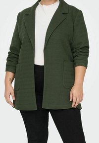 Zelený blazer v herringbone vzoru s klopovým límcem, dvěma předními kapsami a ohrnutými rukávy, v kombinaci s bílým ribbed topem a černými kalhotami.