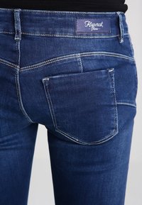 Jeans de mezclilla azul oscuro con sutiles desgastes y costuras en contraste. Cuenta con un parche de marca Kaporal y un bolsillo trasero.